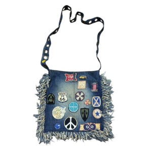 Puede incluir: Bolso de hombro de mezclilla con flecos en la parte inferior y una correa con recortes decorativos. La bolsa está adornada con varios parches, incluyendo un letrero de la Ruta 66, un símbolo de la paz y un logotipo de Miller High Life.