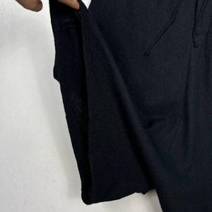 Op de afbeelding: Close-up van een zwart kledingstuk met een trekkoord. De stof lijkt een zacht, getextureerd materiaal te zijn. Het kledingstuk wordt opengehouden, waardoor de binnenkant zichtbaar is. De achtergrond is een effen wit oppervlak.
