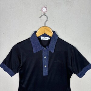 May include: Black polo shirt with a denim collar and sleeve cuffs. The shirt has a three-button placket and the Courr&egrave;ges logo embroidered on the chest. The shirt is hanging on a wooden hanger.