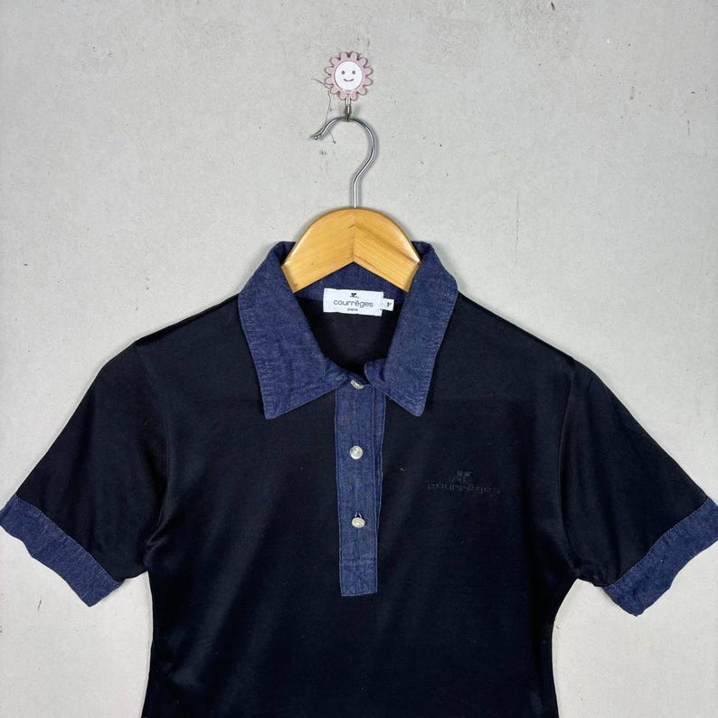 May include: Black polo shirt with a denim collar and sleeve cuffs. The shirt has a three-button placket and the Courr&egrave;ges logo embroidered on the chest. The shirt is hanging on a wooden hanger.