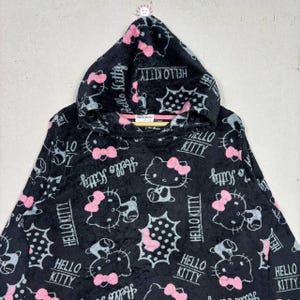 Puede incluir: Sudadera con capucha negra con un patr&oacute;n repetido de personajes de Hello Kitty con lazos rosas y las palabras "Hello Kitty" en blanco. La sudadera tiene una textura suave y afelpada.