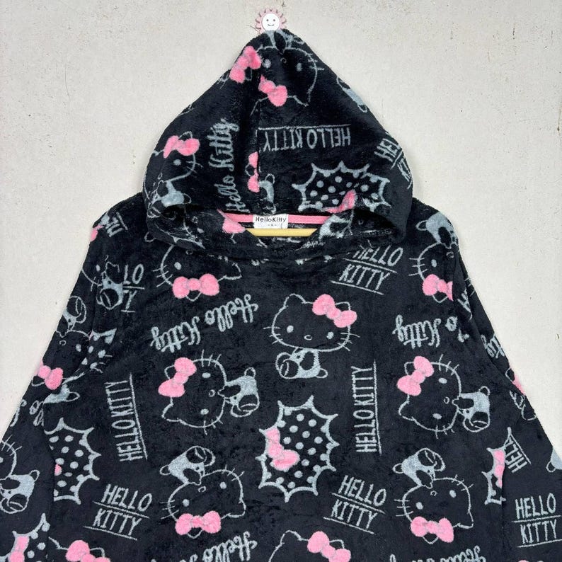 Puede incluir: Sudadera con capucha negra con un patr&oacute;n repetido de personajes de Hello Kitty con lazos rosas y las palabras "Hello Kitty" en blanco. La sudadera tiene una textura suave y afelpada.