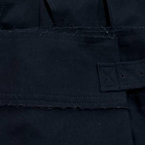 May include: Close-up of a black garment with a rectangular pocket and a belt with a silver buckle. The fabric has a slightly distressed, frayed edge. The belt is secured with a metal buckle.