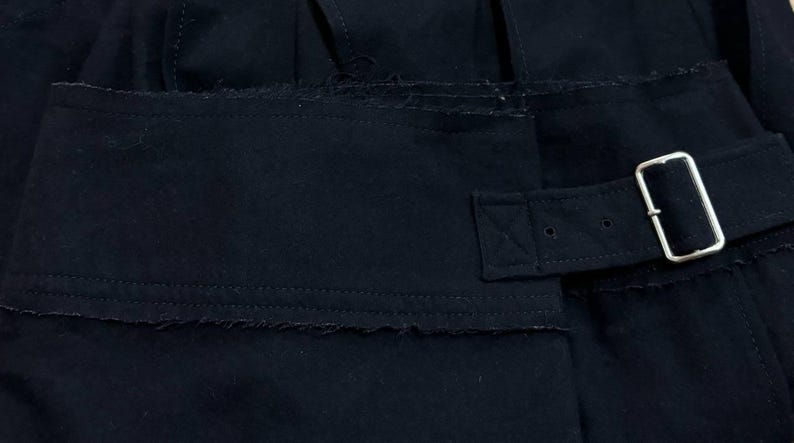 May include: Close-up of a black garment with a rectangular pocket and a belt with a silver buckle. The fabric has a slightly distressed, frayed edge. The belt is secured with a metal buckle.
