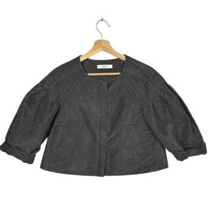 Puede incluir: Chaqueta gris oscuro con mangas tres cuartos colgada de una percha de madera. La chaqueta tiene un cuello redondo y un diseño sencillo sin botones. Las mangas están recogidas en el codo y los hombros tienen un detalle plisado. El nombre de la marca es visible en una pequeña etiqueta.