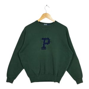 May include: A dark green sweatshirt with a navy blue "P" logo and the word "POLO" embroidered below. The sweatshirt is hanging on a wooden hanger against a white background. The garment has a crew neck and long sleeves.