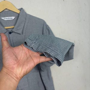 May include: A black and white checkered button-down shirt with a collar and long sleeves. The shirt is hanging on a wooden hanger. The label reads "Vi for living".