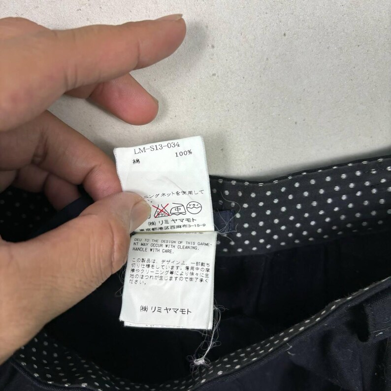 Op de afbeelding: Close-up van een marineblauw kledingstuk met een witte stippelvoering. Een wit label met Japanse tekst en de tekst "LM-S13-034" is zichtbaar. Het label bevat ook wasvoorschriften.