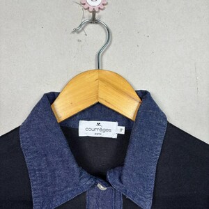 May include: Black shirt with a dark blue denim collar. The shirt has a button closure and a small white label with the text "courr&egrave;ges paris". The shirt is hanging on a wooden hanger.