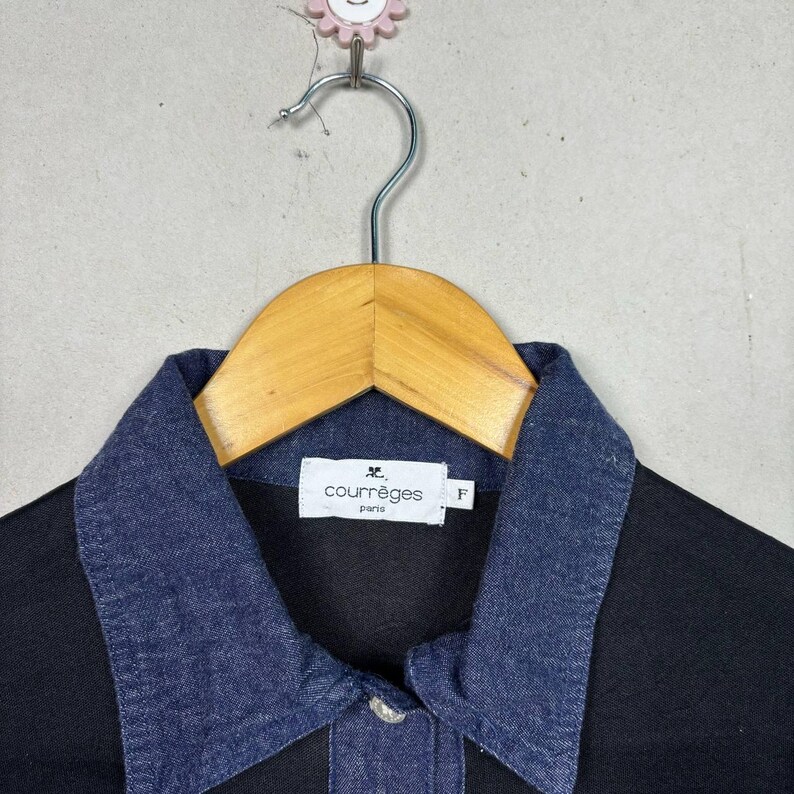 May include: Black shirt with a dark blue denim collar. The shirt has a button closure and a small white label with the text "courr&egrave;ges paris". The shirt is hanging on a wooden hanger.