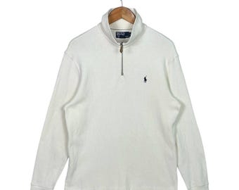 May include: A white long-sleeve shirt with a quarter-zip collar. The shirt features a small navy blue logo on the chest. The shirt is hanging on a wooden hanger.