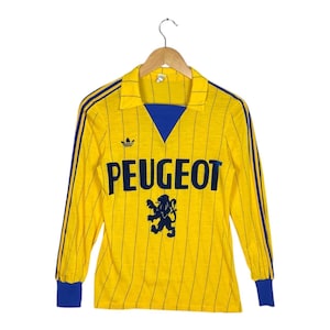 May include: Yellow long-sleeve jersey with blue accents and the word "PEUGEOT" in navy blue. The jersey features the Adidas logo and a lion emblem. The sleeves and collar are trimmed with blue. The jersey is hanging on a wooden hanger.