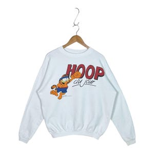 May include: A white long-sleeve sweatshirt featuring a graphic of the orange cartoon cat Garfield wearing a blue headband and blue shorts, jumping to shoot a basketball. The text HOOP du jour is printed in red and black above the character.