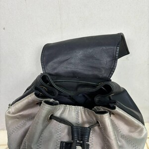 Op de afbeelding: Een zwarte leren rugzak met een zilver-grijze stoffen onderkant. De tas heeft een trekkoordsluiting en een klep die opent om de binnenkant te onthullen. De stiksels zijn zichtbaar en de tas lijkt gemaakt van duurzame materialen.