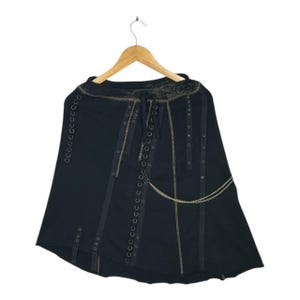 May include: Black skirt with a unique design, hanging on a wooden hanger. The skirt features grommet details, decorative stitching, and a chain accent. The skirt has a layered asymmetrical hemline.