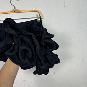 Op de afbeelding: Zwarte shorts met een groot, geplooid stoffen detail. De shorts hangen aan een houten hanger. De geplooide stof voegt een unieke textuur en designelement toe aan het kledingstuk. De achtergrond is neutraal.