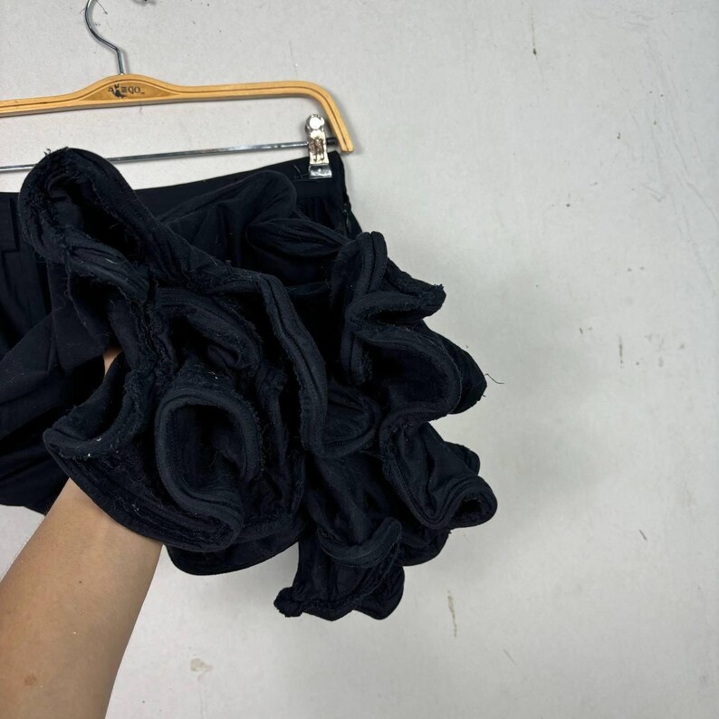 Op de afbeelding: Zwarte shorts met een groot, geplooid stoffen detail. De shorts hangen aan een houten hanger. De geplooide stof voegt een unieke textuur en designelement toe aan het kledingstuk. De achtergrond is neutraal.