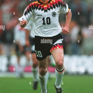 May include: A footballer wearing a white jersey with the number 10, black shorts, and white socks. The jersey has a black, red, and yellow design on the shoulders. The Adidas logo is visible. The player is on a green pitch.