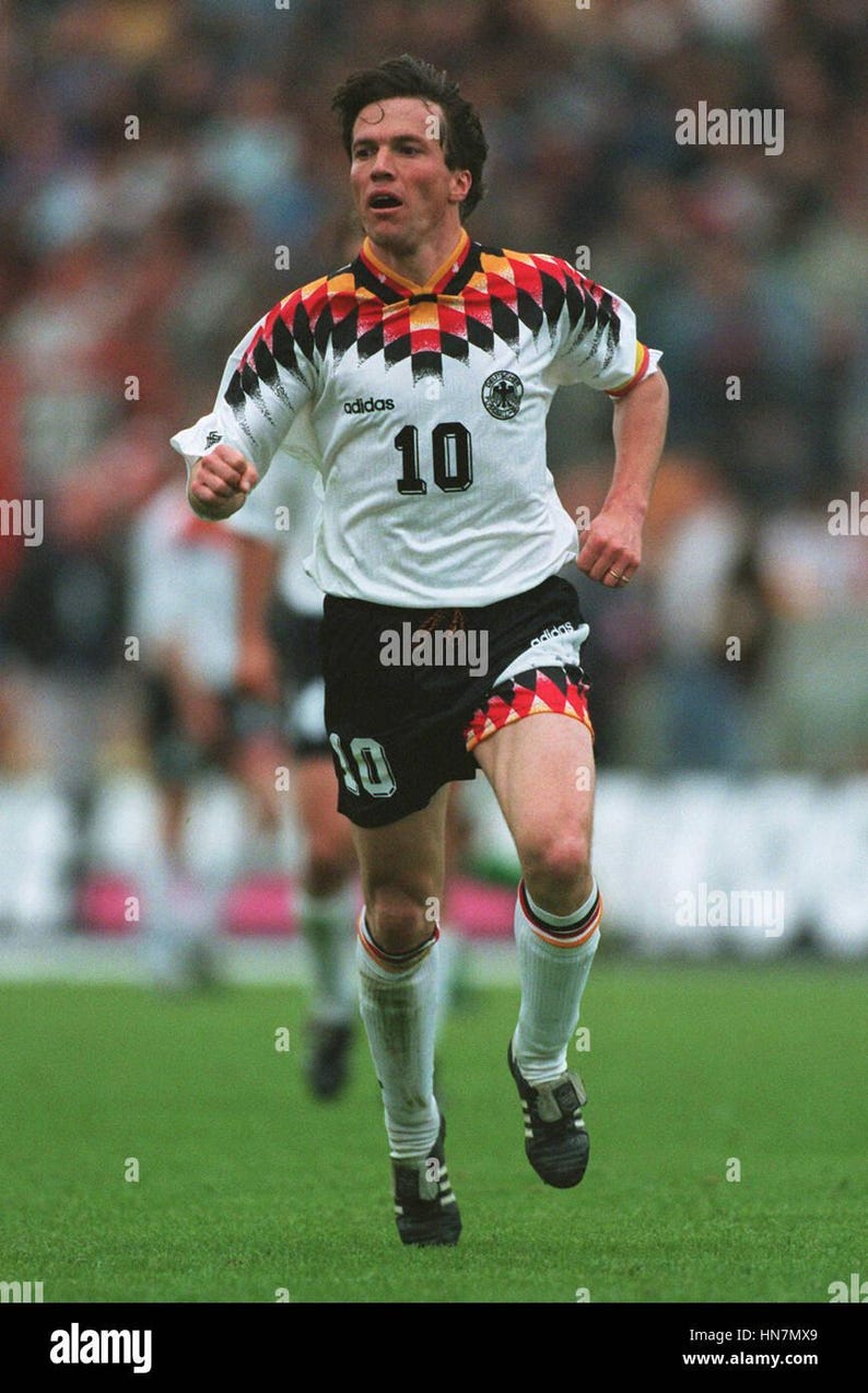 May include: A footballer wearing a white jersey with the number 10, black shorts, and white socks. The jersey has a black, red, and yellow design on the shoulders. The Adidas logo is visible. The player is on a green pitch.