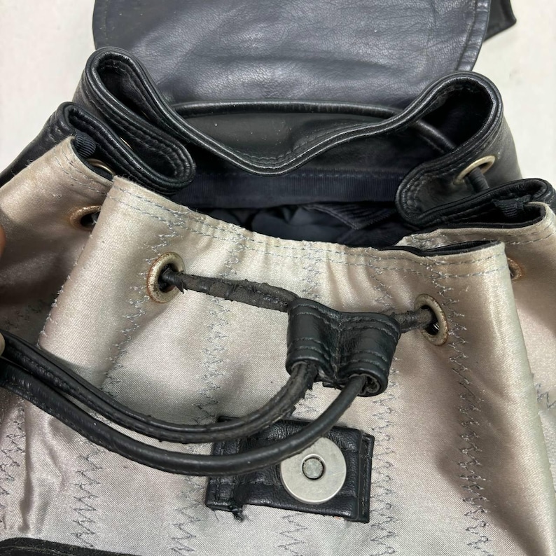 Op de afbeelding: Zwarte leren rugzak met een trekkoordsluiting en zilverkleurige metalen oogjes. De binnenkant is een lichtgrijze stof met zichtbare stiksels. De tas heeft een magnetische druksluiting en zwarte leren banden.