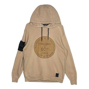 Peut inclure: Sweat à capuche beige avec capuche à cordon et un grand emblème circulaire sur le devant. L'emblème porte l'inscription "BOY LONDON" et un motif d'aigle. Un patch noir est sur la manche gauche.