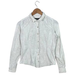 May include: A white button-down shirt with long sleeves, hanging on a wooden hanger. The shirt has a subtle textured pattern and a classic collar. The buttons are small and round. The shirt is a neutral color, suitable for various outfits.