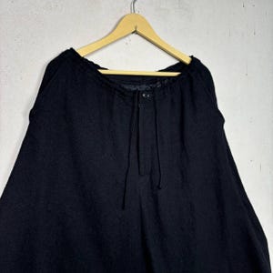Op de afbeelding: Zwarte wollen broek met een tailleband met trekkoord en knoopsluiting, hangend aan een houten hanger. De broek heeft een wijd beenontwerp en een textuurstof.