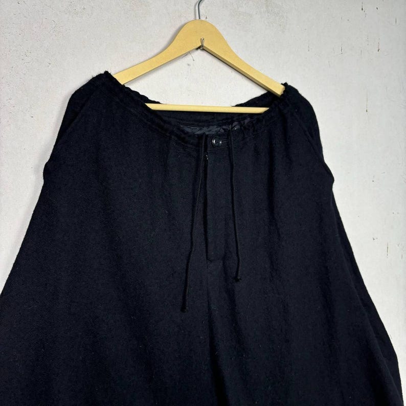 Op de afbeelding: Zwarte wollen broek met een tailleband met trekkoord en knoopsluiting, hangend aan een houten hanger. De broek heeft een wijd beenontwerp en een textuurstof.
