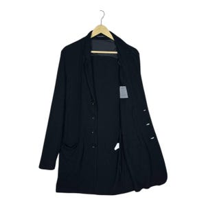 Może przedstawiać: Czarny długi płaszcz z zapięciem na guziki z przodu i dwiema przednimi kieszeniami. Płaszcz wisi na drewnianym wieszaku na białym tle. Płaszcz jest otwarty, odsłaniając podszewkę i małą metkę.