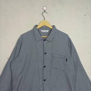 May include: A button-down shirt with a small checkered pattern in black and white, hanging on a wooden hanger. The shirt has a single pocket and the text "Vi for Living" is visible on the collar.