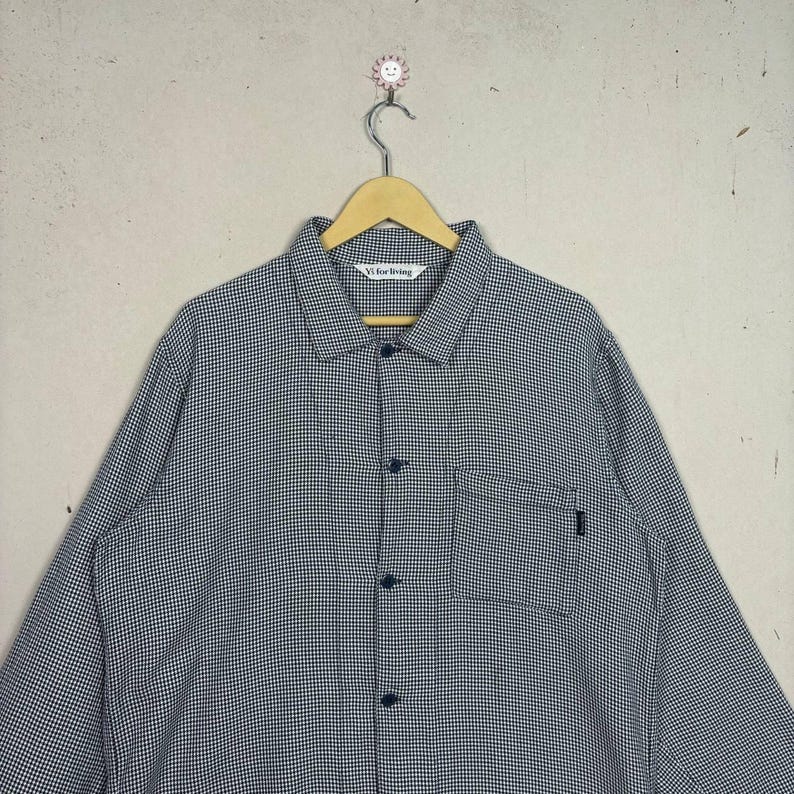 May include: A button-down shirt with a small checkered pattern in black and white, hanging on a wooden hanger. The shirt has a single pocket and the text "Vi for Living" is visible on the collar.