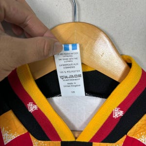 May include: A close-up of a football jersey with a black, red, and yellow design. The jersey has a tag with text in English, French, and German. The tag reads "100% Polyester" and "Made in United Kingdom".