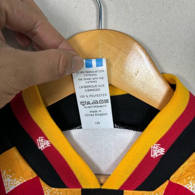 May include: A close-up of a football jersey with a black, red, and yellow design. The jersey has a tag with text in English, French, and German. The tag reads "100% Polyester" and "Made in United Kingdom".