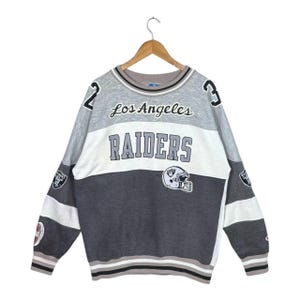 Könnte beinhalten: Ein graues, weißes und schwarzes Los Angeles Raiders Sweatshirt. Das Sweatshirt zeigt den Teamnamen und das Logo, mit der Nummer 2 auf der linken Schulter und der Nummer 3 auf der rechten Schulter. Die Ärmel haben Team-Patches.