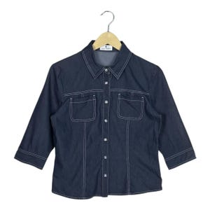 May include: Dark blue denim shirt with three-quarter sleeves, white stitching, and two front pockets. The shirt has a collared neckline and is button-down. The shirt is hanging on a wooden hanger.