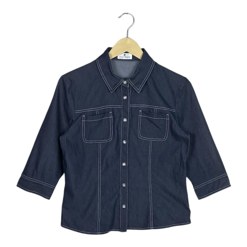 May include: Dark blue denim shirt with three-quarter sleeves, white stitching, and two front pockets. The shirt has a collared neckline and is button-down. The shirt is hanging on a wooden hanger.