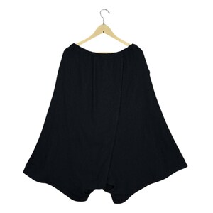 Op de afbeelding: Zwarte broek met wijde pijpen die aan een houten hanger hangt. De broek heeft een uniek, asymmetrisch ontwerp met een wikkelvoorkant. De stof lijkt een zacht, vloeiend materiaal te zijn, geschikt voor vrijetijdskleding.