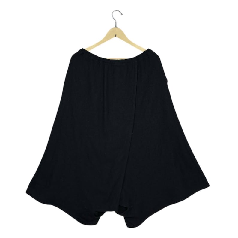 Op de afbeelding: Zwarte broek met wijde pijpen die aan een houten hanger hangt. De broek heeft een uniek, asymmetrisch ontwerp met een wikkelvoorkant. De stof lijkt een zacht, vloeiend materiaal te zijn, geschikt voor vrijetijdskleding.