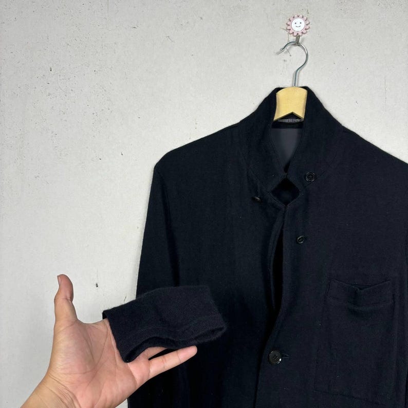 Może przedstawiać: Czarna kurtka z kołnierzem mandaryńskim i zapięciem na guziki. Kurtka ma jedną kieszeń na piersi i złożony mankiet. Kurtka wisi na drewnianym wieszaku na jasnoszarym tle.