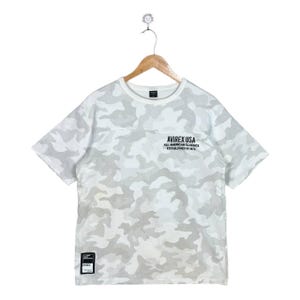 May include: A short-sleeve t-shirt featuring a white and light grey camouflage pattern. The chest displays the black text AVIREX USA ALL AMERICAN CLASSICS ESTABLISHED IN 1975. A small black rectangular patch is located at the bottom left hem.