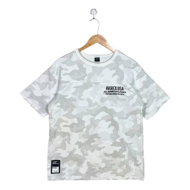 May include: A short-sleeve t-shirt featuring a white and light grey camouflage pattern. The chest displays the black text AVIREX USA ALL AMERICAN CLASSICS ESTABLISHED IN 1975. A small black rectangular patch is located at the bottom left hem.