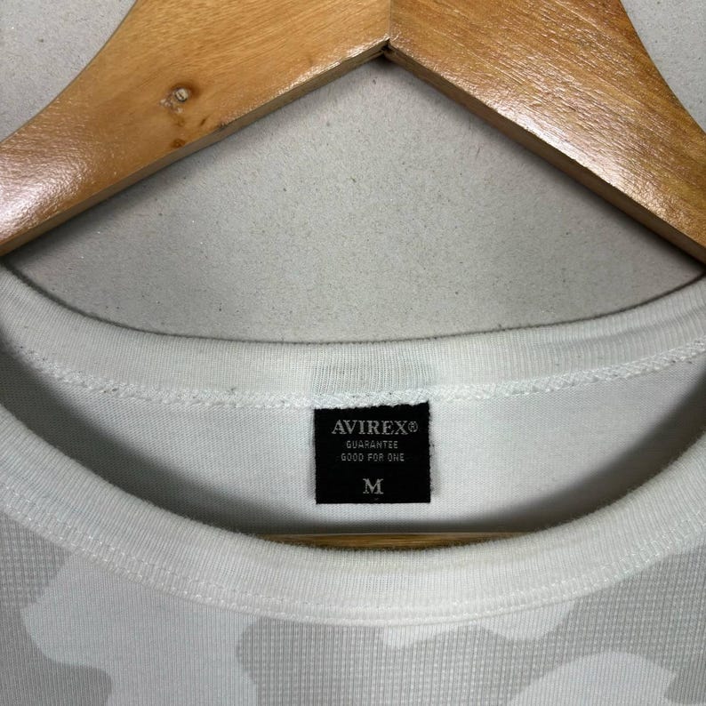 May include: A white t-shirt with a subtle tonal camouflage pattern and a black interior neck label. The label features the text AVIREX, GUARANTEE, GOOD FOR ONE, and the size M. The garment is displayed on a wooden hanger against a neutral background.