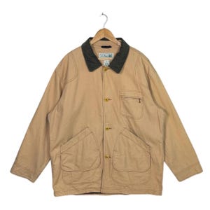 Puede incluir: Chaqueta L.L.Bean color canela con cuello gris oscuro. La chaqueta tiene cuatro bolsillos delanteros, un bolsillo en el pecho con cremallera y cierres de botón. El logotipo de L.L.Bean es visible en el cuello. Cuelga de una percha de madera.
