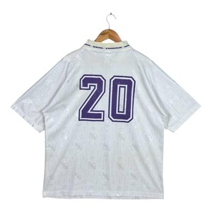 K&ouml;nnte beinhalten: Wei&szlig;es Fu&szlig;balltrikot mit der Nummer 20 in Lila auf dem R&uuml;cken. Das Trikot hat ein strukturiertes Muster und die Worte "REAL MADRID" sind auf dem Kragen aufgedruckt. Das Trikot h&auml;ngt an einem Holzkleiderb&uuml;gel.