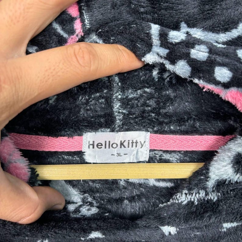 Puede incluir: Primer plano de una sudadera con capucha Hello Kitty negra y rosa. La sudadera tiene una textura difusa con un patr&oacute;n de formas blancas y rosas. La etiqueta dice "Hello Kitty - 3L".