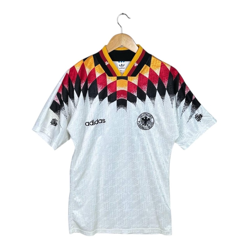 May include: White football shirt with a geometric design in black, red, and yellow on the shoulders and collar. The Adidas logo is on the chest, and a team emblem is on the right. The shirt hangs on a wooden hanger.