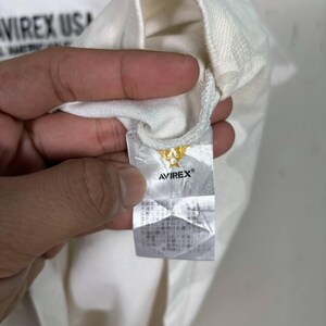 May include: A white garment featuring a black printed logo that reads AVIREX USA. A white fabric care tag is attached to the interior, displaying a gold winged star emblem above the black text AVIREX and Japanese instructional text.