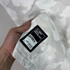 May include: A white and light grey camouflage patterned fabric featuring a rectangular black label with white text that reads U.S. ARMED FORCES MANUFACTURER AVIREX NEW YORK N.Y. EST. 1975, stitched onto the corner of the garment material.