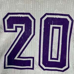 K&ouml;nnte beinhalten: Nahaufnahme eines Sporttrikots mit der Nummer 20 in Lila mit wei&szlig;em Rand. Der Hintergrund ist wei&szlig; mit einem sich wiederholenden Muster aus Text und Logos. Das Trikot ist wahrscheinlich f&uuml;r ein Fu&szlig;ballteam.