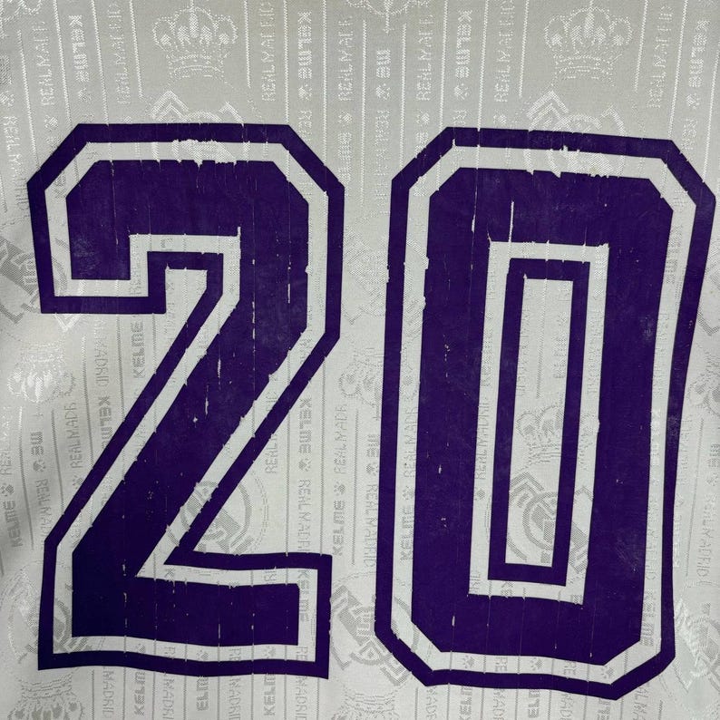 K&ouml;nnte beinhalten: Nahaufnahme eines Sporttrikots mit der Nummer 20 in Lila mit wei&szlig;em Rand. Der Hintergrund ist wei&szlig; mit einem sich wiederholenden Muster aus Text und Logos. Das Trikot ist wahrscheinlich f&uuml;r ein Fu&szlig;ballteam.